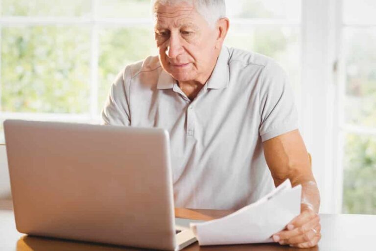 Senior-man-holding-papers-and-looking-at-laptop