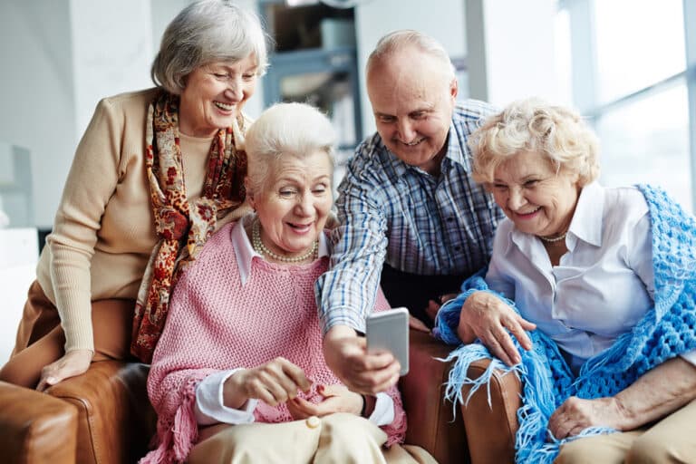 Senior man showing phone to 3 senior women, all laughing and smiling